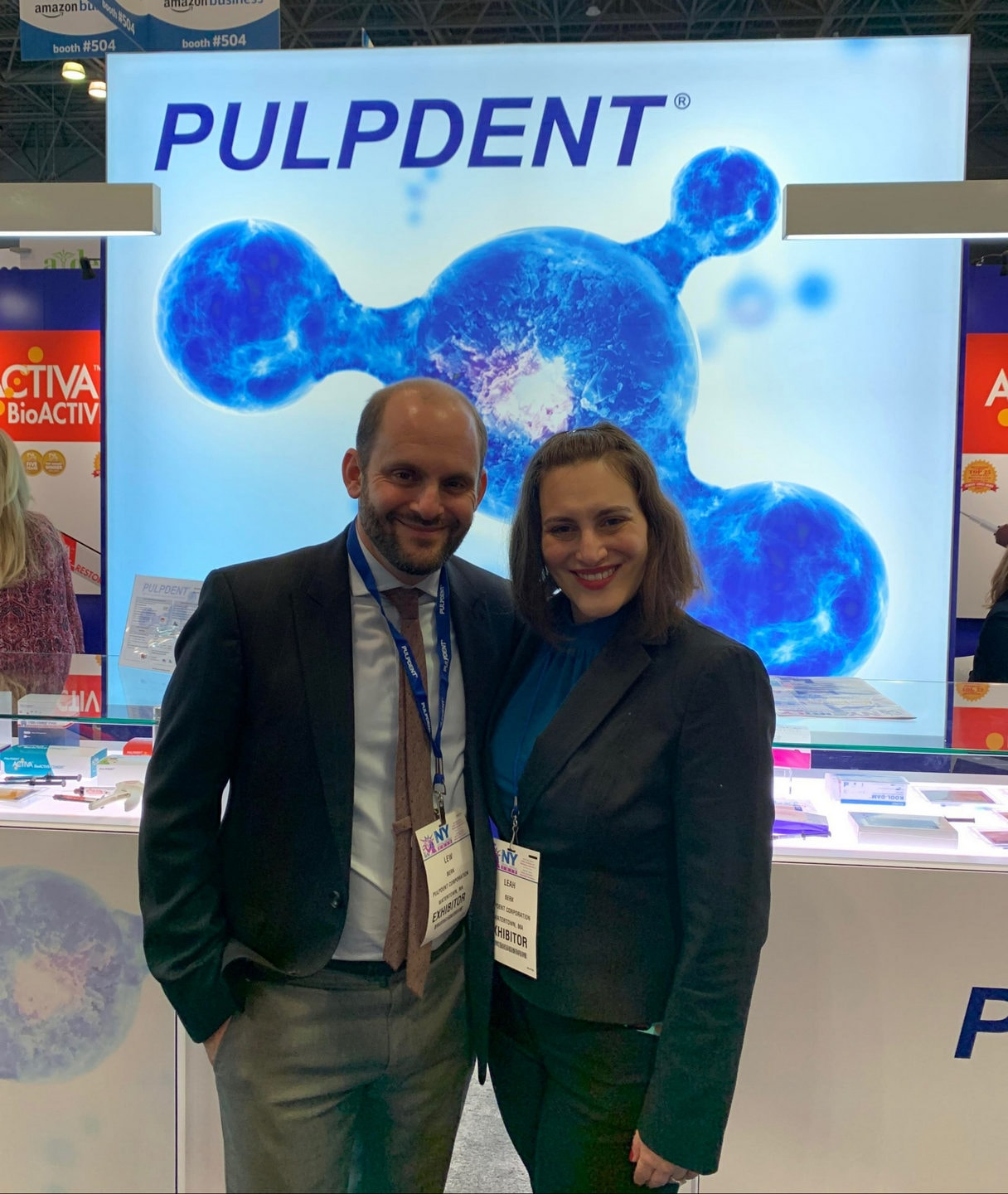 Lewis Berk and Leah Berk stand facing and smiling at the camera at a trade show. Behind them is a graphic representing an ion with the words Pulpdent right above it.