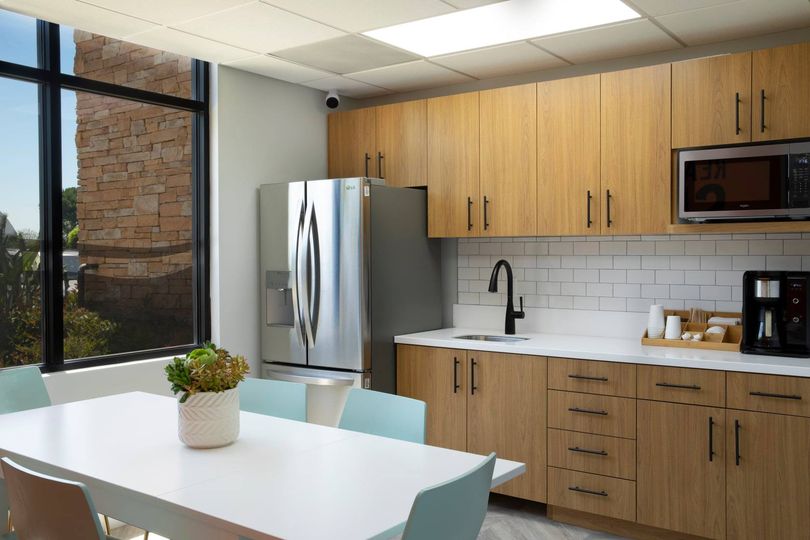 A workplace kitchen featuring white tables and modern wood cabinets, a sink, silver fridge, and microwave.