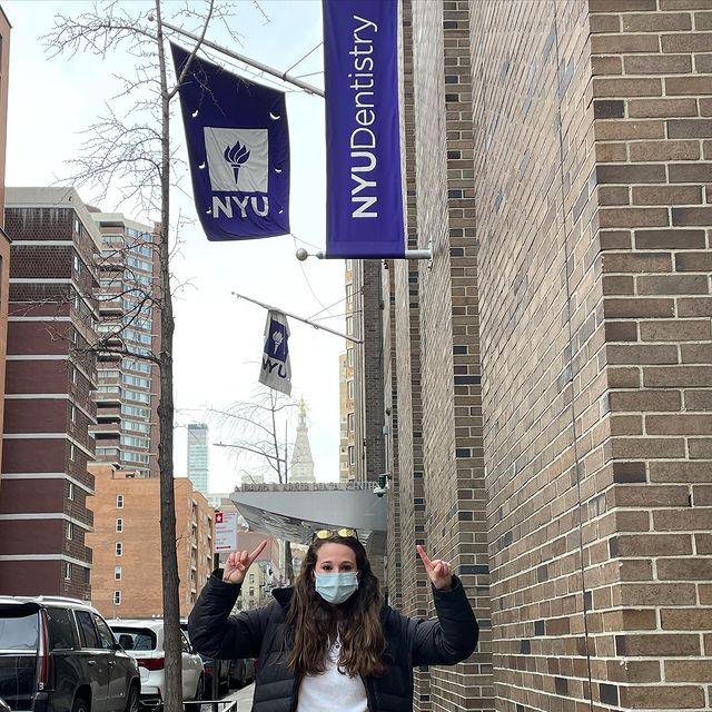 Whitney Lynn First Year Dental Student at NYU pointing to NYU flag above her outside the school