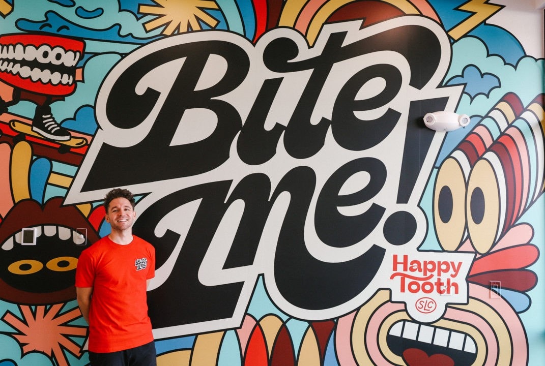 Dr. Tyler Hanks is wearing a red shirt. He is standing in front of a large, colorful mural that prominently features the words "Bite Me."
