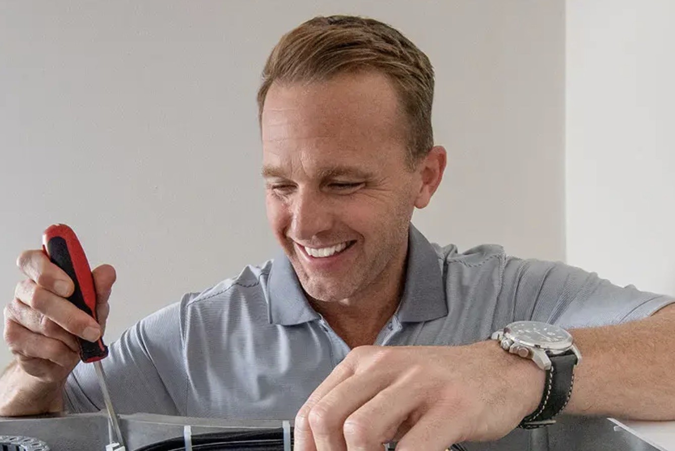 A smiling man in a blue polo uses a screwdriver to fix a bundle of cables in place.