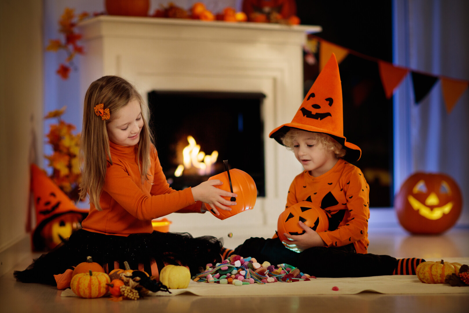A young girl and boy dressed for Halloween dump their buckets of candy into a large pile on the floor between them.