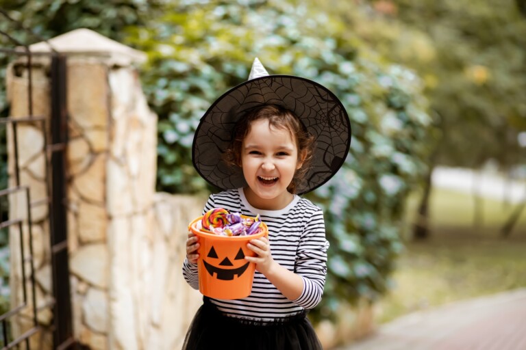 A young girl in a witch hat stands on the sidewalk, smiling, with a pumpkin-shaped container of candy.