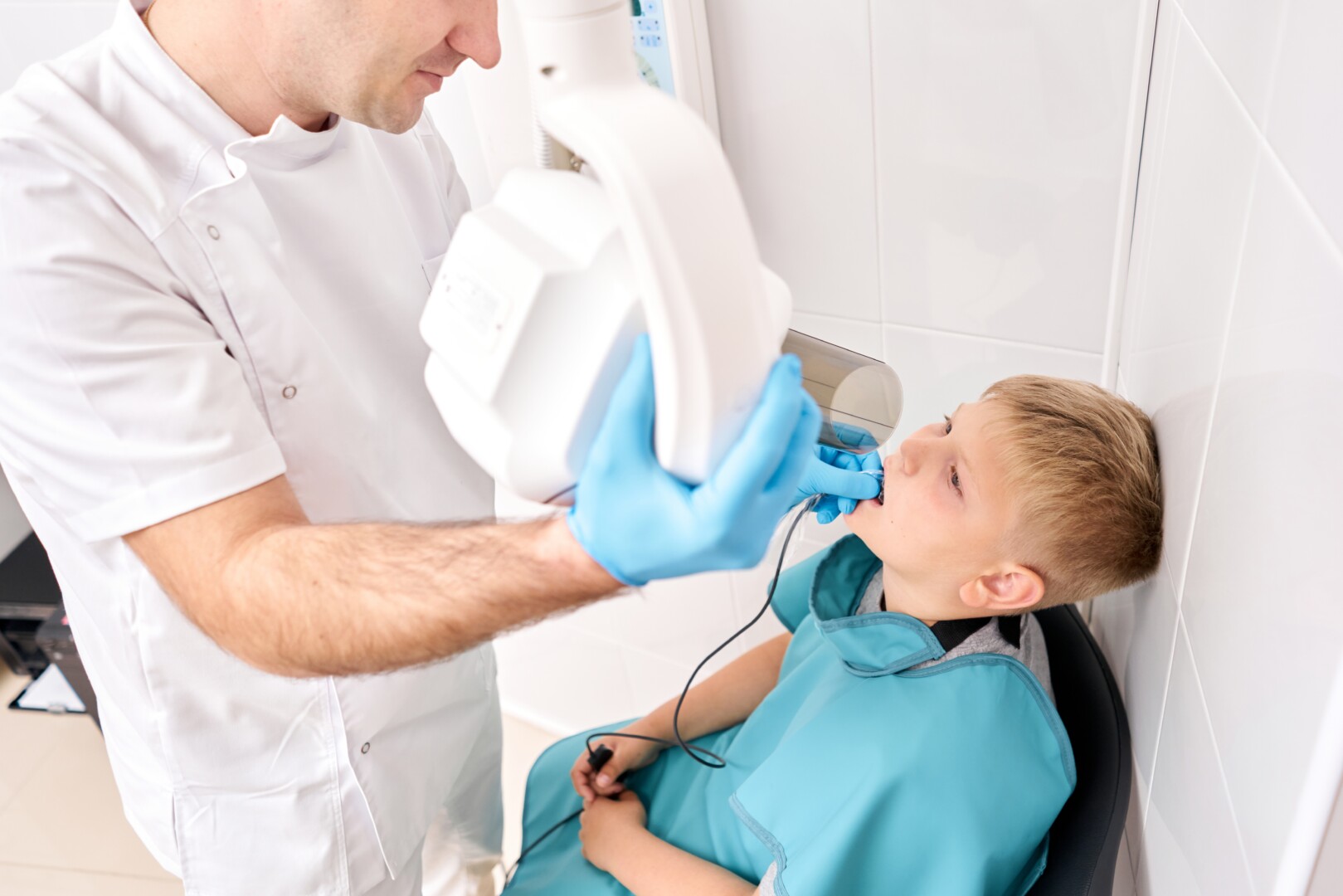 A dentist places an x-ray camera in a child's mouth to take images of the child's teeth. The child wears a light blue lead apron for protection.