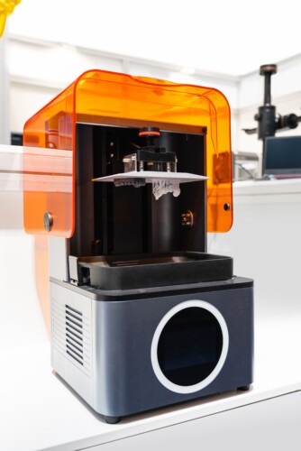 A 3D printer sits with its orange shield open to reveal the device's interior.