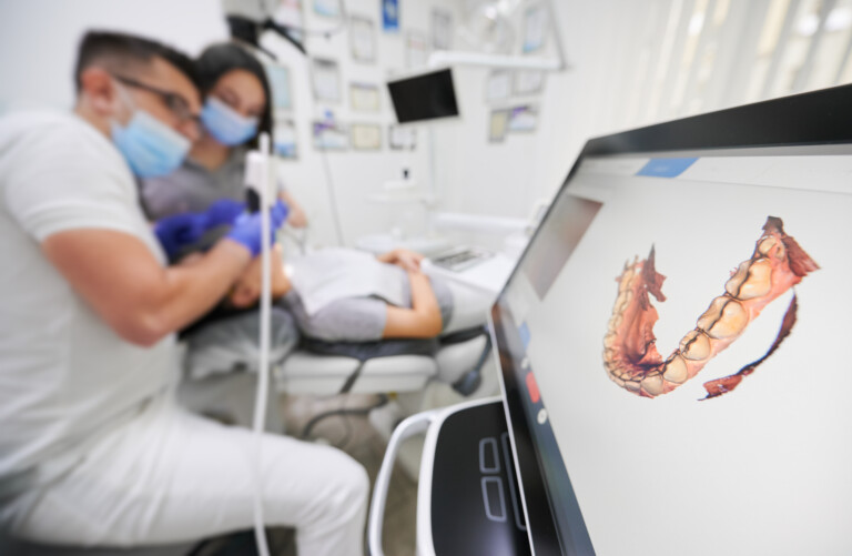 In the background, a dentist uses a digital scanning tool that is creating an image of the patient's mouth on a screen in the foreground.