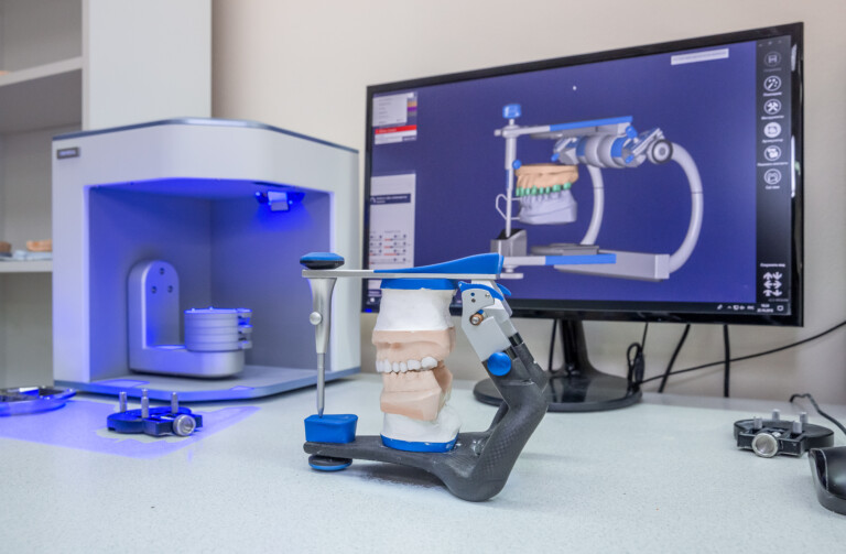 A set of 3D printed dentures sit held by the machine's housing. A digital image of the dentures is on a computer screen behind the dentures. The 3D printer sits to the left. It is open and is glowing blue.