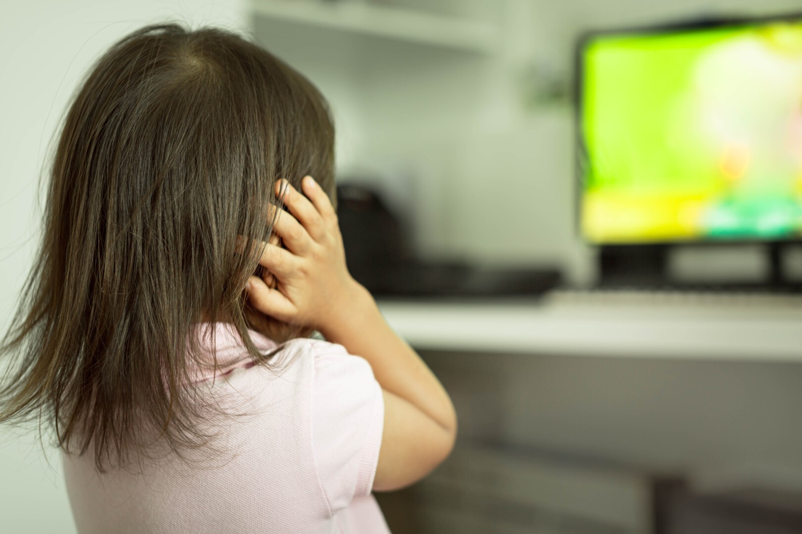 A young girl with sensory processing disorder facing away from the camera covers her ears with her hands. A television is on in the background of the image.
