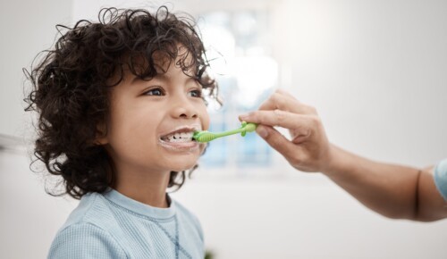 A child with curly hair is exposing their teeth while an adult hand from the right of the frame brushes their teeth for them.