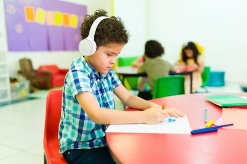An autistic child with curly hair wears large white headphones to block out the sound of the surrounding classroom.