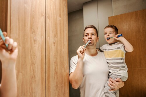 A father holds his child while the two of them brush their teeth while watching themselves in the bathroom mirror.
