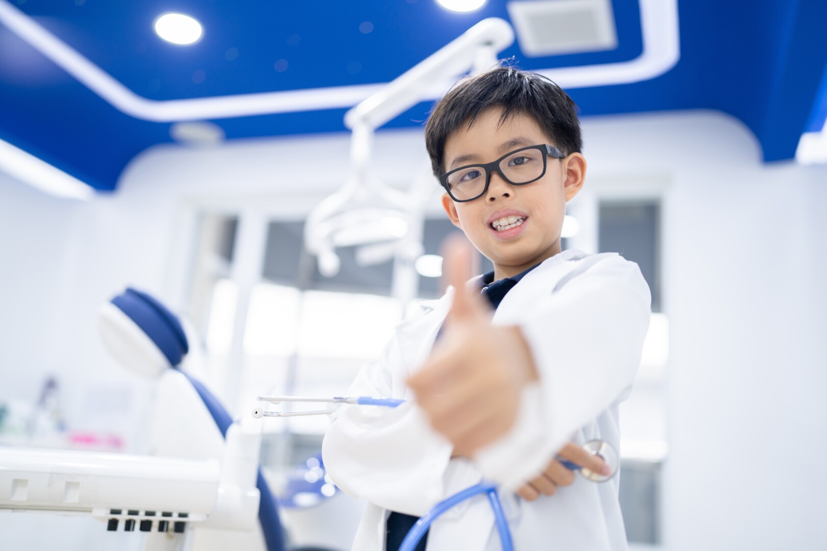 A young boy in glasses and a medical coat in a dental office aims a thumbs up at the camera.