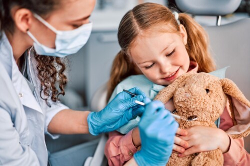 A dentist in a white mask and blue gloves demonstrates proper brushing technique on a small girl's stuffed rabbit.