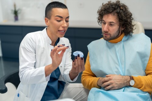 A female doctor with a buzzcut (left) holds a dental model while explaining something to a patient (right), a curly haired male.