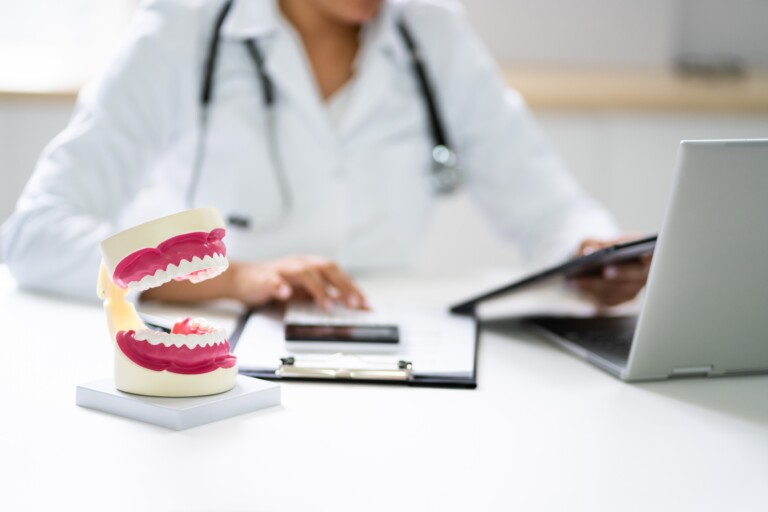 A red model of teeth sits on the table in the foreground. In the background, a dentist works with a laptop and a calculator.